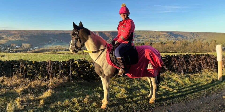 Person sitting on a horse in a rural landscape, wearing a pink hat and matching coat. The horse has a pink covering. Stone wall and hills in the background under a clear blue sky.